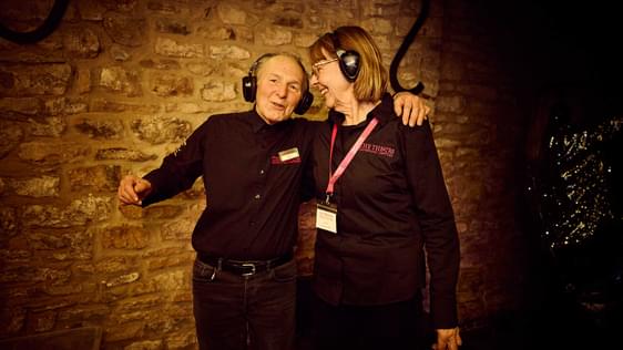 Two ushers in black shirts with pink Theatre logos have their arms round each other, smiling