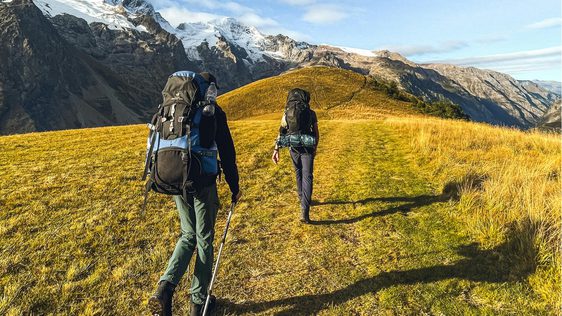 Two hikers walking across along a grass path towards a snow capped peak.