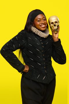 Efé is a black woman with straight black hair. She wears a black Elizabethan style jacket with a ruff and smiles whilst holding a skull.