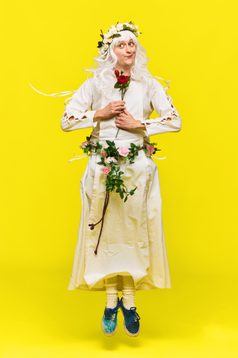 Tom is a white man wearing a white dress and wig. He is holding a red rose and jumping in the air. His wig and waist are adorned with flowers.