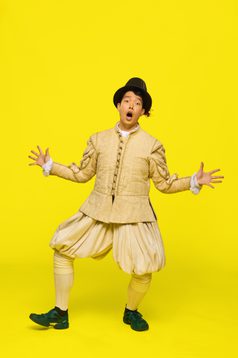 Woogie is a South Korean man with black short hair. He is wearing a black top hat with a beige Elizabethan style jacket and bloomers. He has is arms outstretched and looks surprised with his mouth open wide.