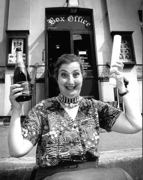 Tamara Malcom sits outside The Theatre holding a bottle of champagne and a scroll of paper, looks to be celebrating.