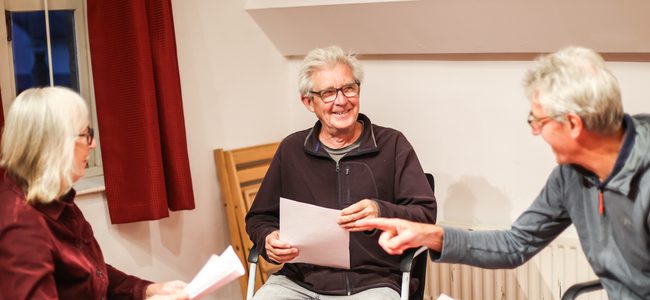 Two men and a woman sit and discuss a script. They are smiling.