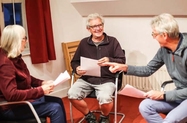 Two men and a woman sit and discuss a script. They are smiling.