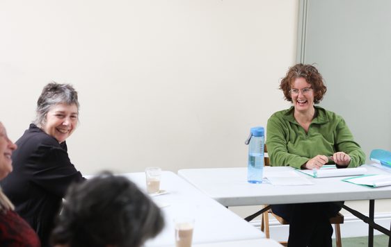 A woman in green sits and smiles across the table at others. Next to her another woman with short grey hair is smiling.