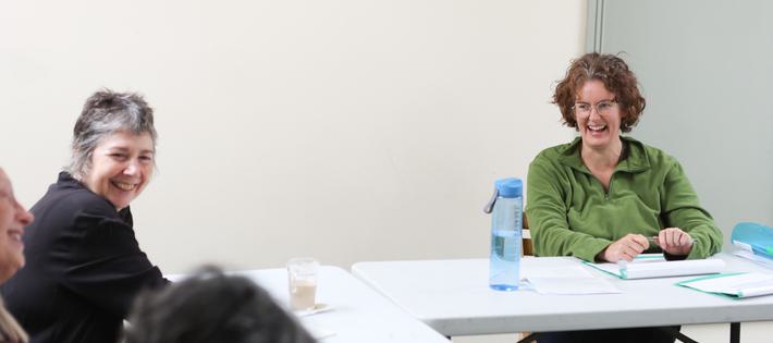 A woman in green sits and smiles across the table at others. Next to her another woman with short grey hair is smiling.