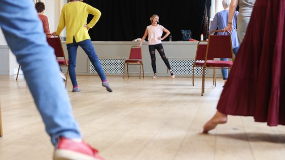 A group practice ballet exercises, pointing their leg and foot out to the side leg by a dance teacher