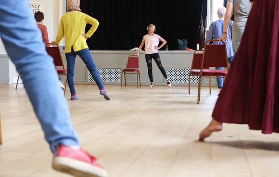 A group practice ballet exercises, pointing their leg and foot out to the side leg by a dance teacher