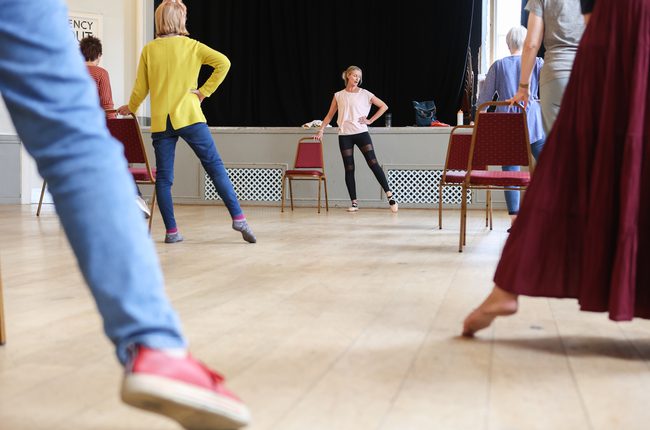 A group practice ballet exercises, pointing their leg and foot out to the side leg by a dance teacher