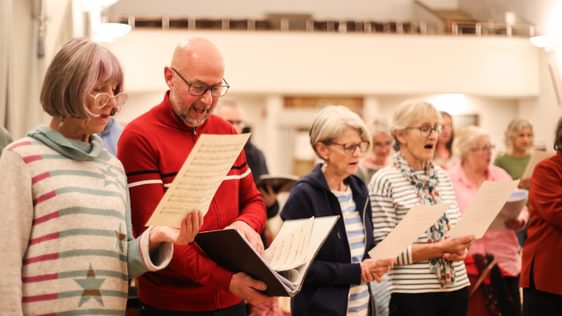 A group of people sing together, reading sheet music and lyrics held in front of them.