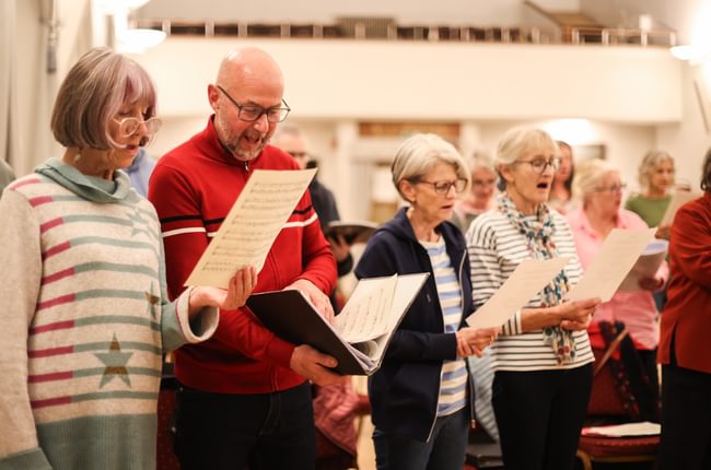 A group of people sing together, reading sheet music and lyrics held in front of them.