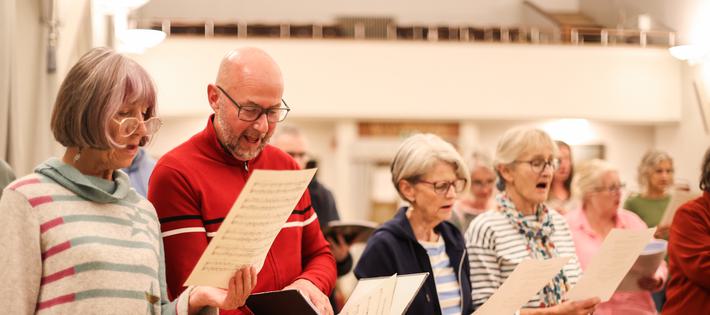 A group of people sing together, reading sheet music and lyrics held in front of them.