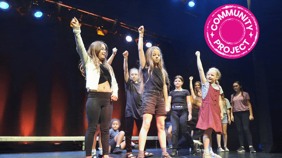 Group of participants performing on stage with their right hand punching up in the air with Community Project Badge