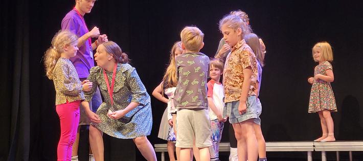 A group of small children rehearsing onstage. The teacher is acting like a monkey to make a small girl laugh.