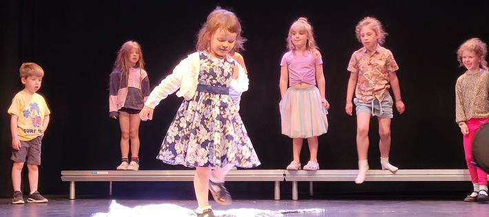 A group of small children are performing onstage. The little girl in the foreground of the picture is wearing a floral dress and is jumping on a large piece of foil sheet, pretending that it is a puddle.
