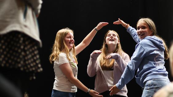Two teen girls use their arms to create a circle in the air. A third girl pokes her head through it, her arms up as she acts like a dog. All three are laughing.