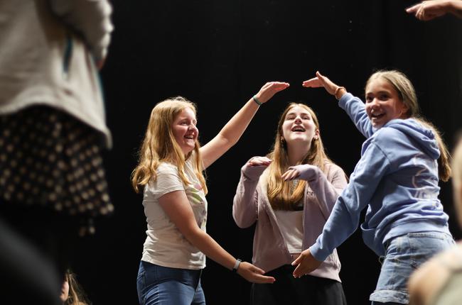 Two teen girls use their arms to create a circle in the air. A third girl pokes her head through it, her arms up as she acts like a dog. All three are laughing.