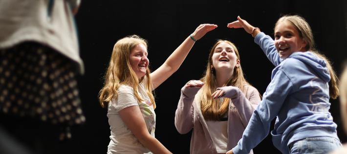 Two teen girls use their arms to create a circle in the air. A third girl pokes her head through it, her arms up as she acts like a dog. All three are laughing.