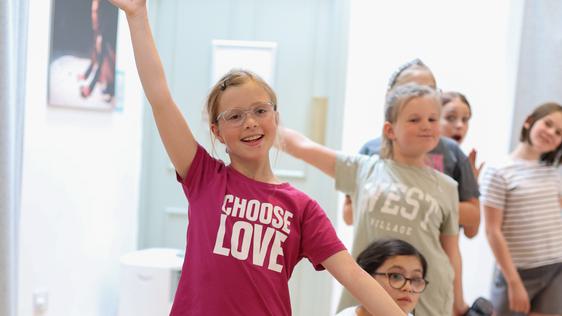 A young girl in a pink 'choose love' t-shirt poses as though she has just finished dancing. Behind her, several other children strike different end poses.