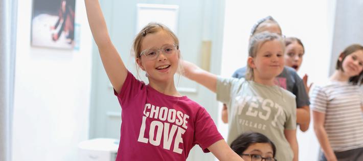 A young girl in a pink 'choose love' t-shirt poses as though she has just finished dancing. Behind her, several other children strike different end poses.