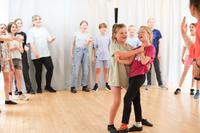 Two girls are in the centre of the room, as though they have just completed a spin mid-dance. Around the edge of the room, other children watch.