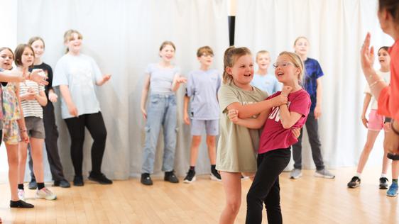 Two girls are in the centre of the room, as though they have just completed a spin mid-dance. Around the edge of the room, other children watch.