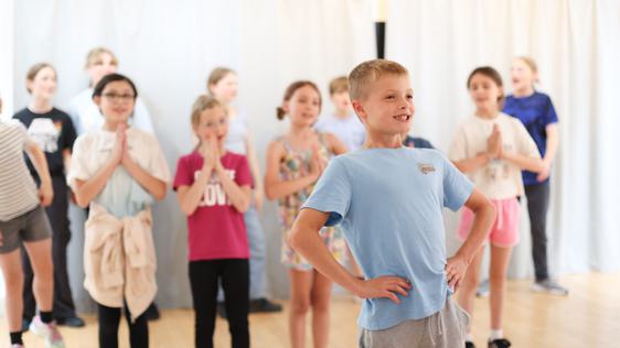 A boy stands confident in the foreground. He is wearing a pale blue t-shirt and has his hands on his hips. Behind him are a row of girls with their hands in a prayer position.