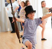 A boy is knelt on one knee with his arms out in a 'ta-da!' motion. He wears a top hat and is holding an umbrella.