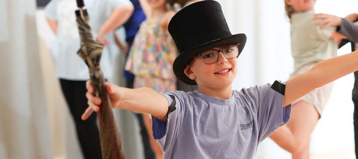 A boy is knelt on one knee with his arms out in a 'ta-da!' motion. He wears a top hat and is holding an umbrella.