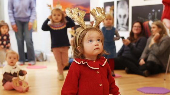 A tiny girl in a red cardigan is wearing sparkly gold reindeer antlers. She looks up.