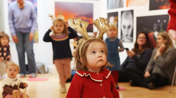A tiny girl in a red cardigan is wearing sparkly gold reindeer antlers. She looks up.