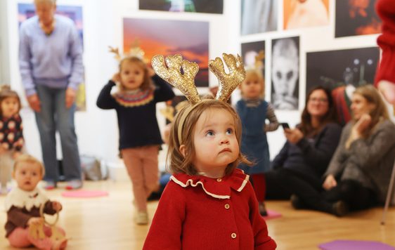 A tiny girl in a red cardigan is wearing sparkly gold reindeer antlers. She looks up.
