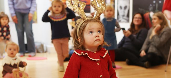 A tiny girl in a red cardigan is wearing sparkly gold reindeer antlers. She looks up.