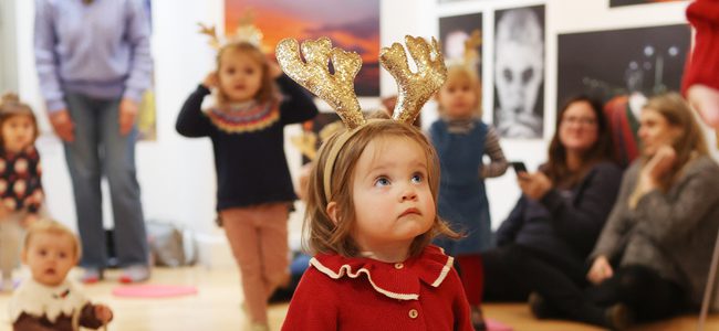 A tiny girl in a red cardigan is wearing sparkly gold reindeer antlers. She looks up.