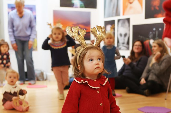 A tiny girl in a red cardigan is wearing sparkly gold reindeer antlers. She looks up.