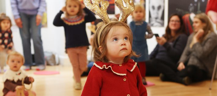 A tiny girl in a red cardigan is wearing sparkly gold reindeer antlers. She looks up.
