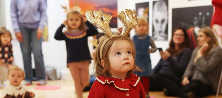 A tiny girl in a red cardigan is wearing sparkly gold reindeer antlers. She looks up.