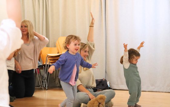 A small girl in a purple cardigan is running, her arms out like wings. Behind her, a mother and her young daughter are stretching their arms up to the ceiling as part of a drama exercise.