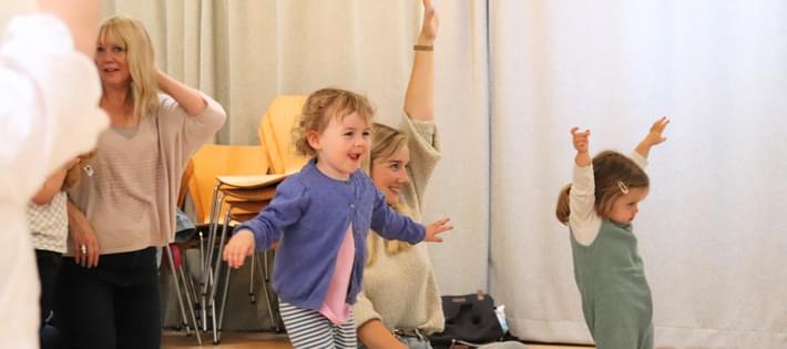 A small girl in a purple cardigan is running, her arms out like wings. Behind her, a mother and her young daughter are stretching their arms up to the ceiling as part of a drama exercise.