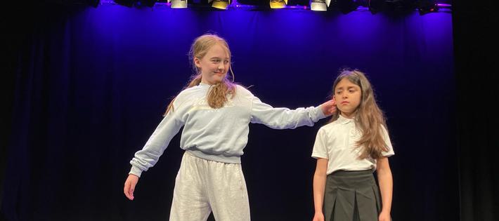 Two girls stand on stage. One of them is acting as a mean character, and is grabbing the other girl's ear.