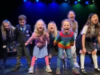 A group of children are performing on stage; they stand with powerful stances. They stick their tongues out at the camera in a playful and defiant way.