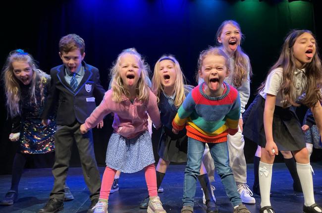 A group of children are performing on stage; they stand with powerful stances. They stick their tongues out at the camera in a playful and defiant way.
