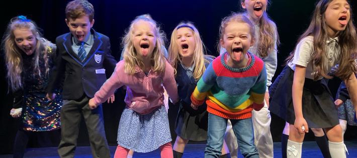 A group of children are performing on stage; they stand with powerful stances. They stick their tongues out at the camera in a playful and defiant way.