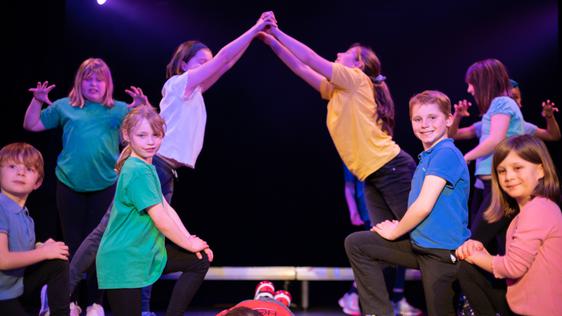 A group of children wearing colourful shirts perform on stage. Two of them create an archway with their hands.
