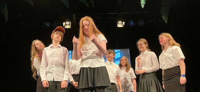 A group of teenagers in white shirts are on stage. A girl in the centre is reading something that causes intrigue.