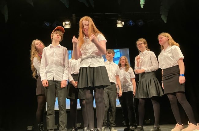 A group of teenagers in white shirts are on stage. A girl in the centre is reading something that causes intrigue.