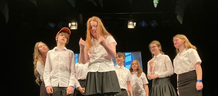 A group of teenagers in white shirts are on stage. A girl in the centre is reading something that causes intrigue.