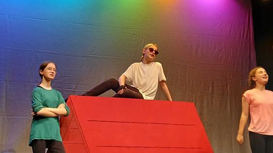 A teen boy sits on top of a giant red dog kennel, dressed as Snoopy. On either side of him girls dressed in green and pink react to what he says. There are multi-coloured lights in the background.