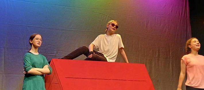 A teen boy sits on top of a giant red dog kennel, dressed as Snoopy. On either side of him girls dressed in green and pink react to what he says. There are multi-coloured lights in the background.
