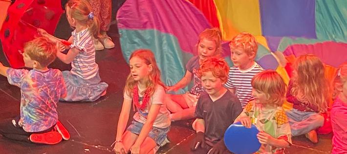 A group of small children on stage using a colourful parachute, umbrellas and frisbee to create the image of a car - Chitty Chitty Bang Bang.
